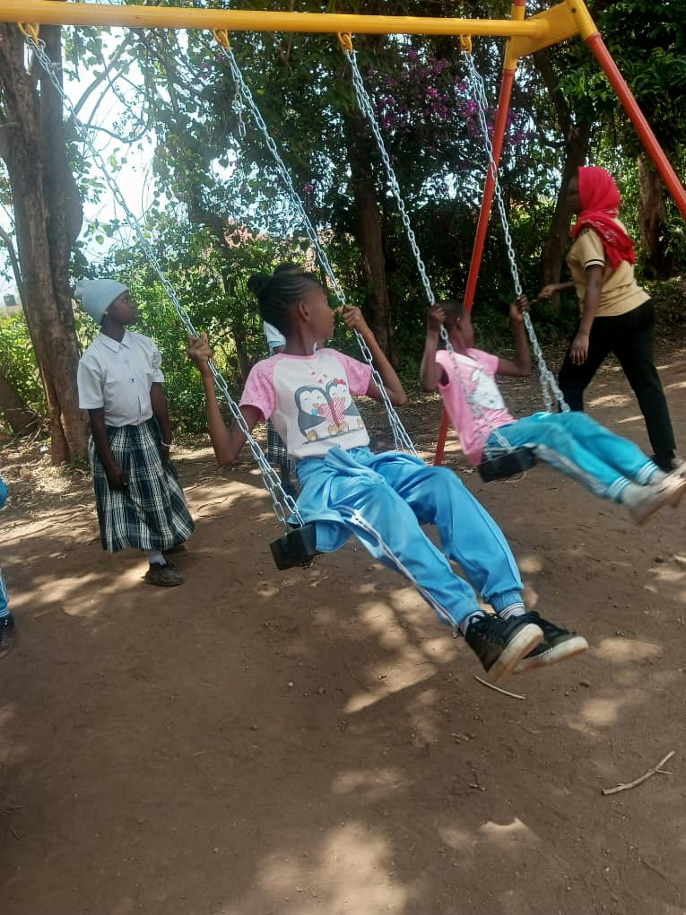 Two girls are sitting on swings, while two other girls stand nearby on a dirt playground with trees and greenery in the background.