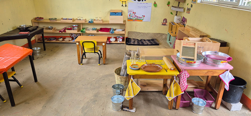 A classroom with small tables and chairs, shelves holding various utensils and containers, and baskets on the floor. Bright decorations and learning materials are visible on the walls and shelves.