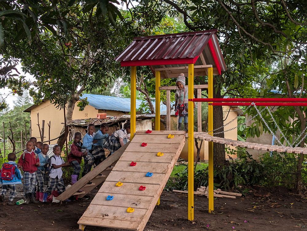 Children gather near a small playground structure with a wooden climbing wall and roof, surrounded by trees and a building in the background.