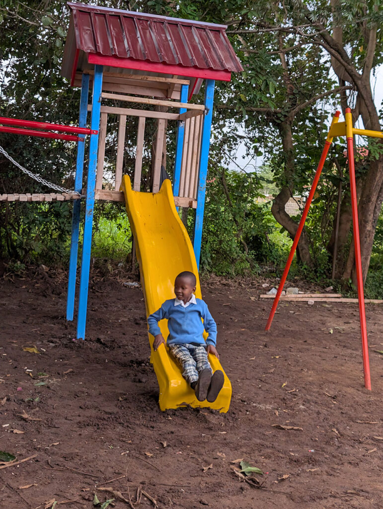 A child in a blue sweater slides down a yellow slide on a playground surrounded by trees and dirt.
