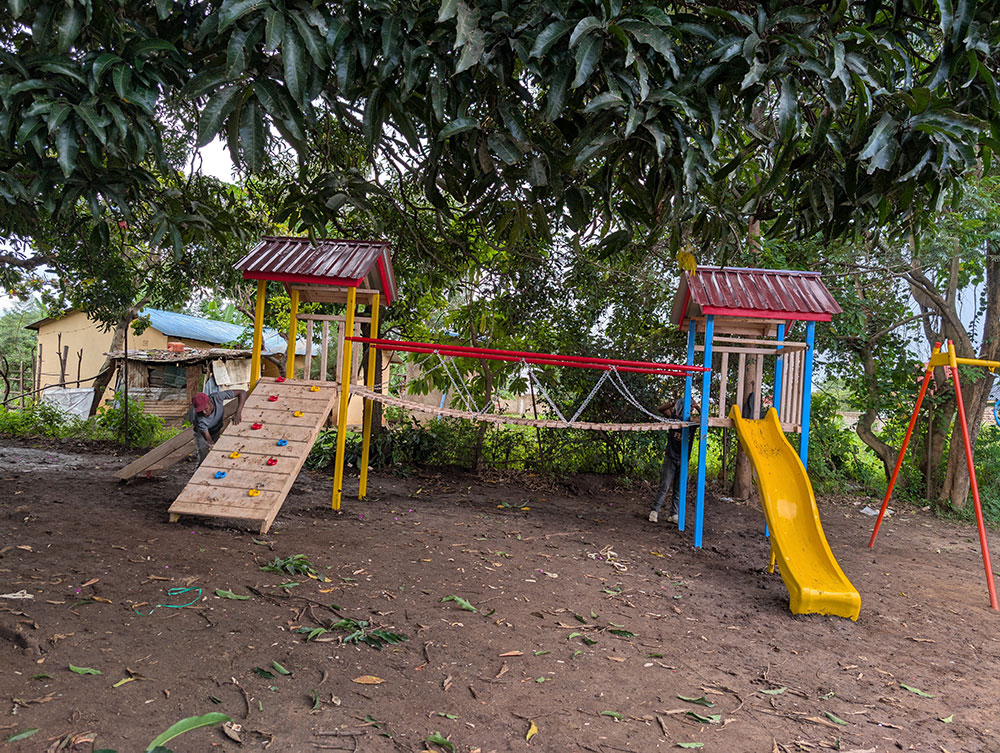 Children’s outdoor playset with a yellow slide, a climbing wall, two small roofs, and a rope bridge, set on bare ground under large trees.