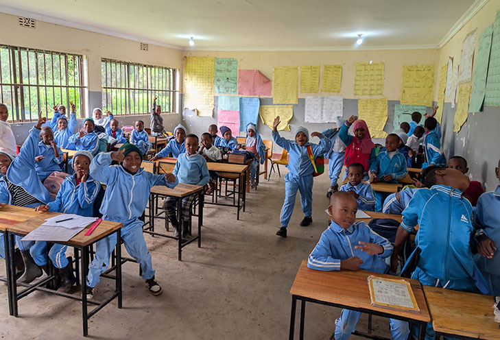A classroom with many students in blue uniforms sitting and standing at desks, some raising their hands, with educational posters on the walls.