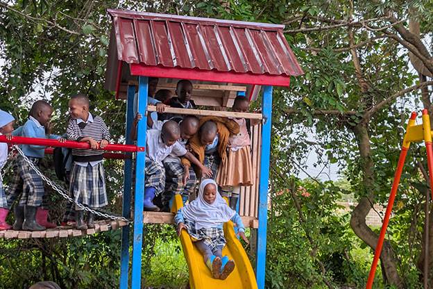 Several children in school uniforms play on a playground structure; one girl slides down a yellow slide while others watch from above. Trees and greenery surround the play area.