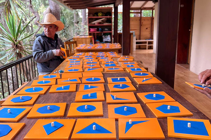 A person wearing a hat sits at a table covered with orange boards displaying various blue geometric shapes, in an indoor-outdoor setting.