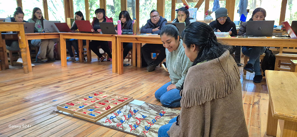 Two women sit on the floor arranging letters on a wooden board while a group of people with laptops observe from tables in a wood-paneled room.