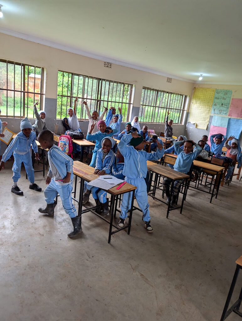 A classroom with children in blue uniforms, some sitting at desks and others standing and interacting, with large windows and posters on the walls.