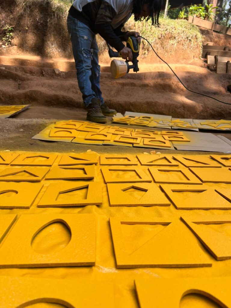 A person uses a spray gun to paint yellow geometric shapes on multiple boards laid out on the ground outdoors.