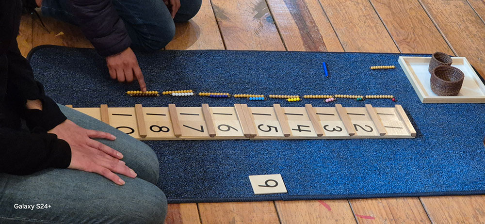 Two people sit on a wooden floor with a blue mat, arranging Montessori math materials including number tiles and bead bars for counting activities.