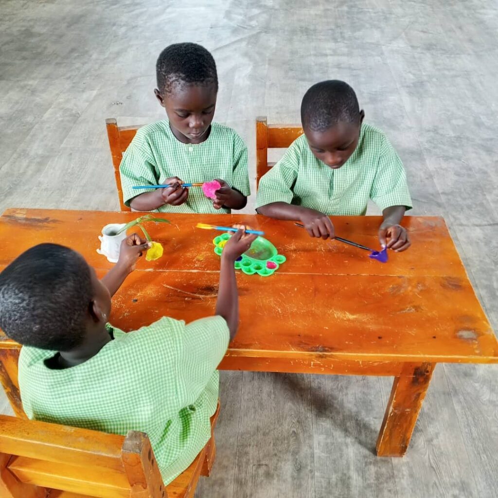Three children in green uniforms sit at a wooden table, painting with brushes and colorful paints on trays in a spacious room with a gray floor.