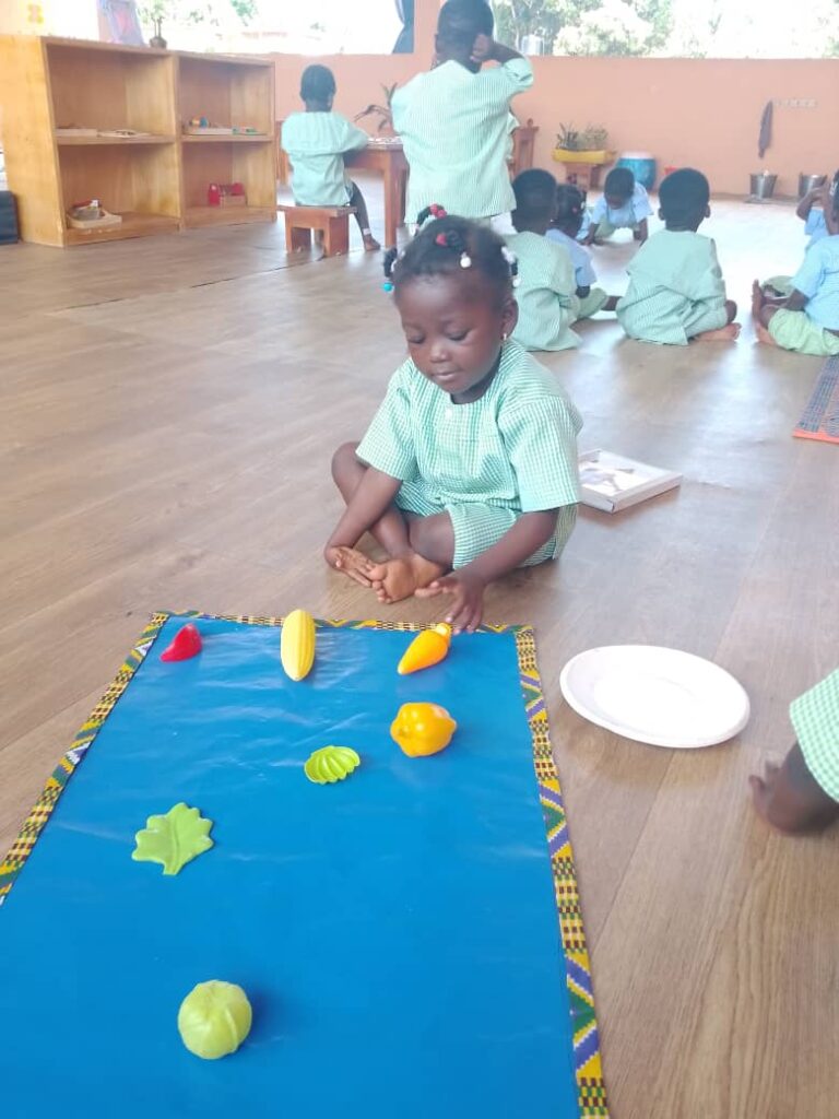 A young girl in a green uniform sits on the floor arranging plastic fruits and vegetables on a blue mat in a classroom with other children in the background.