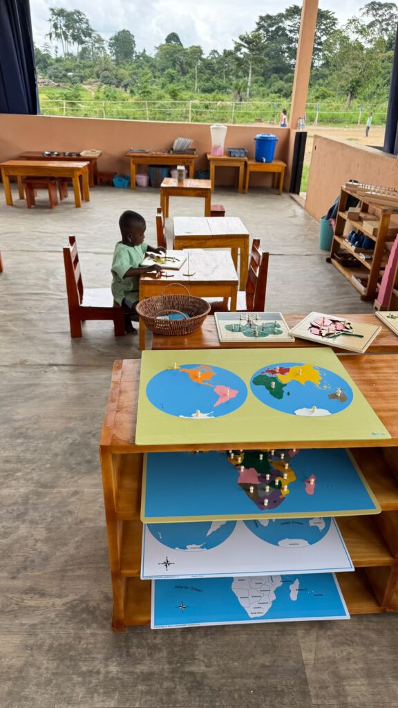 A young child sits at a table in a classroom with educational materials, including world maps and puzzles, on shelves in the foreground. The classroom is open to an outdoor view.