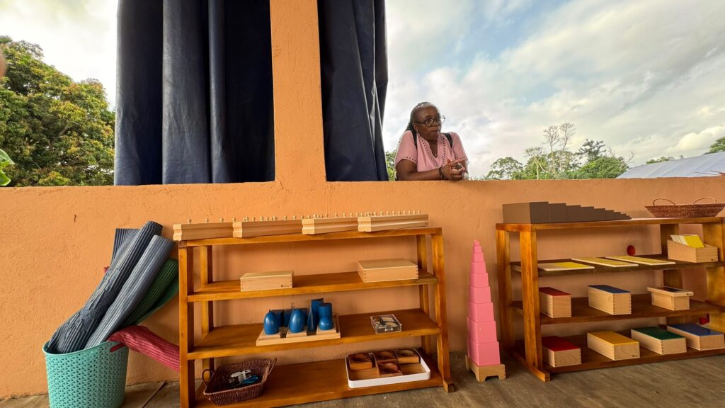 A woman leans on an orange windowsill above wooden shelves holding Montessori educational materials and baskets, with yoga mats and trees visible outside.