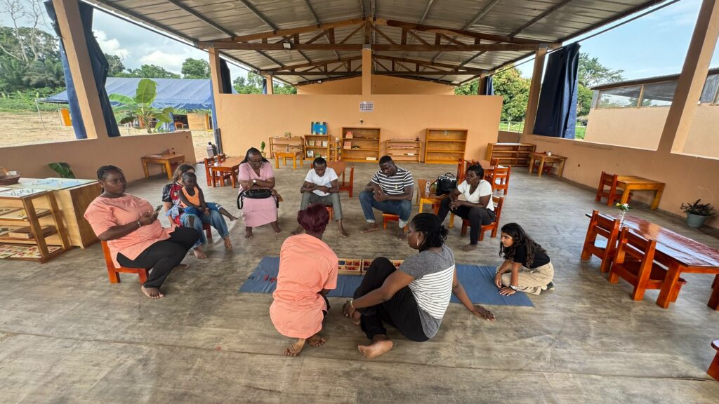 Nine adults sit in a circle on the floor and chairs in a covered open-air classroom, engaging in a group activity with wooden blocks.