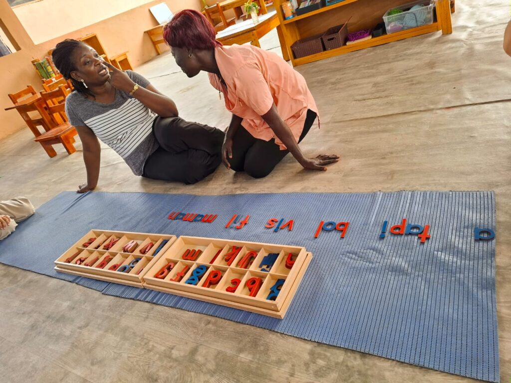 Two women sit on the floor next to a blue mat with Montessori movable alphabet letters arranged and stored in a wooden box.