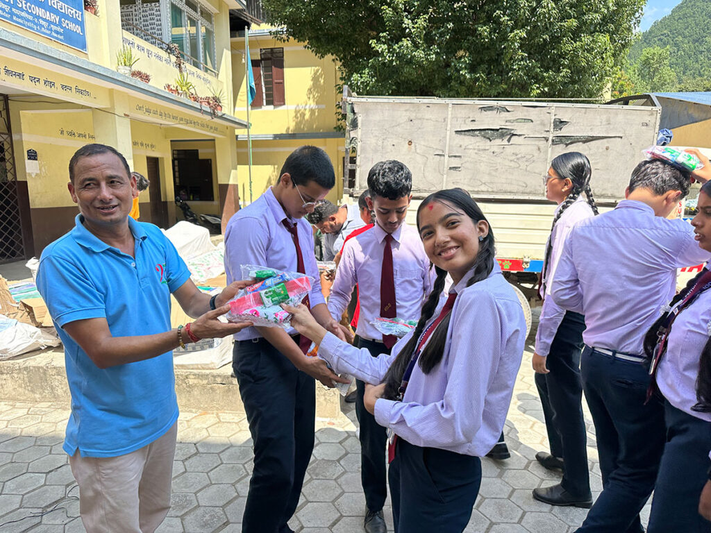 A man in a blue polo shirt hands a package of school supplies to a female student in uniform, surrounded by other students in a school courtyard.