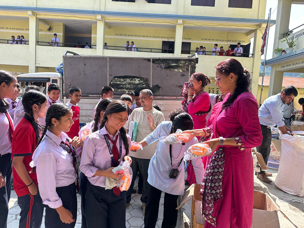 A woman in a pink kurta hands out small plastic bags of snacks to students in school uniforms in an outdoor courtyard.